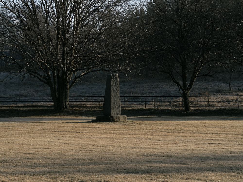 A small weathered stone cenotaph standing alone on frosted grass in a village park, bare trees behind it on a cold overcast morning.